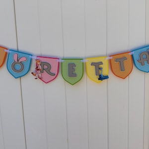 May include: A decorative banner spelling the name LORETTA on individual shield-shaped pennants in orange, blue, pink, green, and yellow. Each pennant features a grey letter and cartoon character accents, all strung together on a white ribbon.