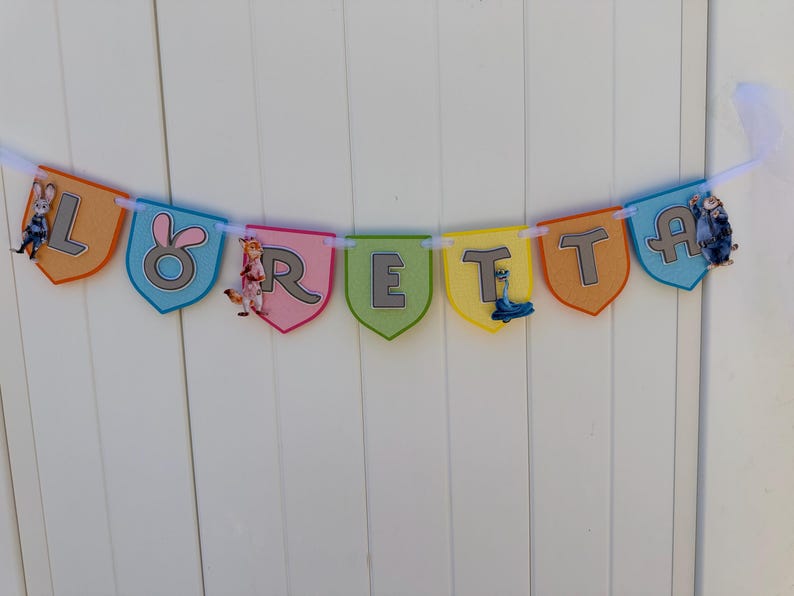 May include: A decorative banner spelling the name LORETTA on individual shield-shaped pennants in orange, blue, pink, green, and yellow. Each pennant features a grey letter and cartoon character accents, all strung together on a white ribbon.