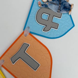 May include: A decorative banner featuring two shield-shaped pennants. The top blue pennant displays a grey letter H and a cartoon cheetah police officer holding a donut. The bottom orange pennant features a grey letter T with a textured surface.