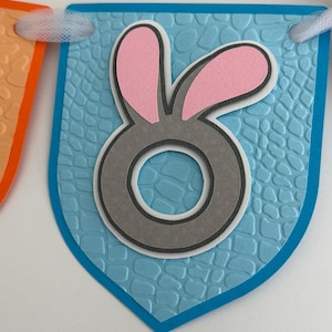 May include: A light blue pennant flag featuring a textured, embossed pattern and a central gray letter O with pink bunny ears attached to the top. The pennant is strung with white tulle ribbon, part of a decorative banner for festive occasions.