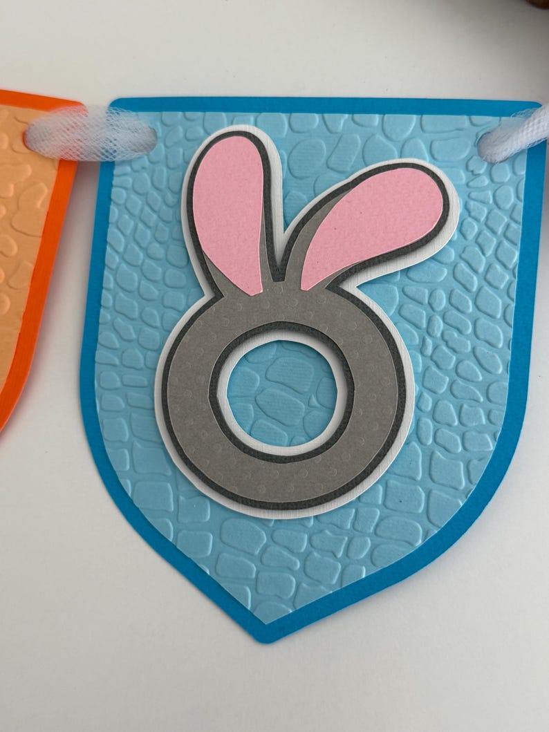 May include: A light blue pennant flag featuring a textured, embossed pattern and a central gray letter O with pink bunny ears attached to the top. The pennant is strung with white tulle ribbon, part of a decorative banner for festive occasions.