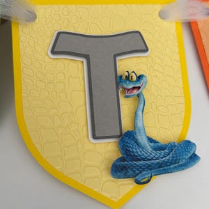 May include: A yellow shield-shaped banner piece featuring a textured reptile-skin pattern, a large gray letter T with a white border, and a blue cartoon snake with wide yellow eyes and an open mouth, attached to a white ribbon at the top corners.