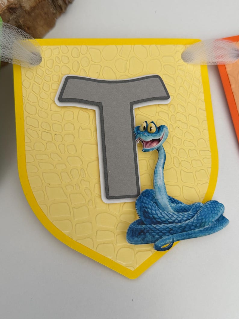 May include: A yellow shield-shaped banner piece featuring a textured reptile-skin pattern, a large gray letter T with a white border, and a blue cartoon snake with wide yellow eyes and an open mouth, attached to a white ribbon at the top corners.