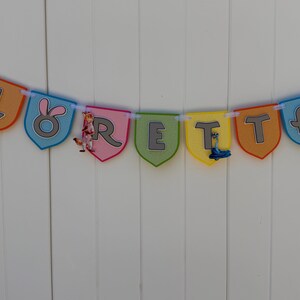 May include: A decorative banner spelling the name LORETTA on individual shield-shaped pennants in orange, blue, pink, green, and yellow. Each pennant features a grey letter and cartoon animal character accents, all strung together on a white ribbon.