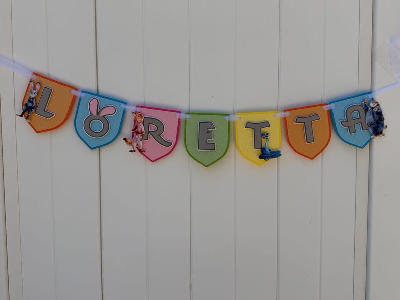 May include: A decorative banner spelling the name LORETTA on individual shield-shaped pennants in orange, blue, pink, green, and yellow. Each pennant features a grey letter and cartoon animal character accents, all strung together on a white ribbon.