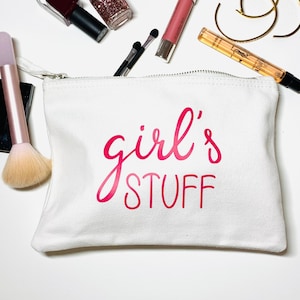 Könnte beinhalten: Weiße Canvas-Kosmetiktasche mit dem Aufdruck "girl's stuff" in Pink.