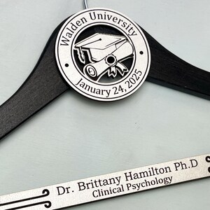 May include: A black wooden hanger with a silver engraved plaque. The plaque features a graduation cap and scroll with the text "Walden University January 24, 2025". A second silver plaque reads "Dr. Brittany Hamilton Ph.D. Clinical Psychology".