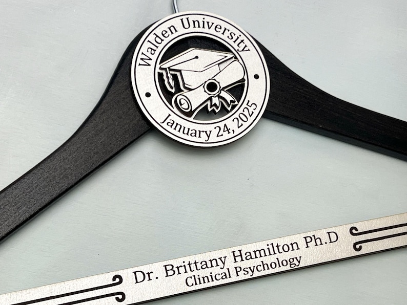 May include: A black wooden hanger with a silver engraved plaque. The plaque features a graduation cap and scroll with the text "Walden University January 24, 2025". A second silver plaque reads "Dr. Brittany Hamilton Ph.D. Clinical Psychology".