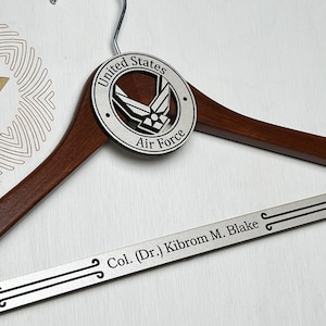 May include: A dark brown wooden clothes hanger featuring a circular silver-toned emblem with the text United States Air Force and the Air Force logo. The bottom bar is silver-toned and engraved with the text Col. (Dr.) Kibrom M. Blake.