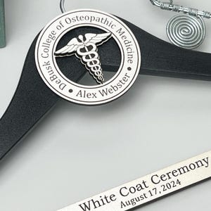 May include: A black wooden clothes hanger featuring a circular silver medallion with a caduceus symbol and the text DeBusk College of Osteopathic Medicine Alex Webster. A silver bar displays White Coat Ceremony August 17, 2024.