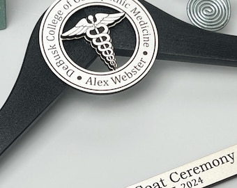 May include: A black wooden clothes hanger featuring a circular silver medallion with a caduceus symbol and the text DeBusk College of Osteopathic Medicine Alex Webster. A silver bar displays White Coat Ceremony August 17, 2024.