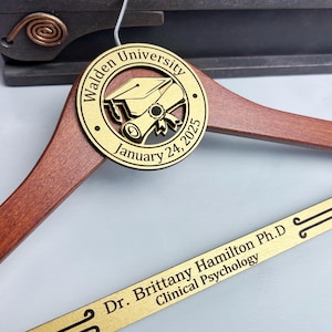 May include: A wooden graduation hanger with a gold-colored round plaque. The plaque has a graduation cap and scroll design with the text "Walden University January 24, 2025". The hanger also has a gold-colored strip with the text "Dr. Brittany Hamilton Ph.D. Clinical Psychology".
