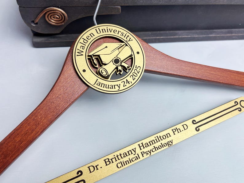 May include: A wooden graduation hanger with a gold-colored round plaque. The plaque has a graduation cap and scroll design with the text "Walden University January 24, 2025". The hanger also has a gold-colored strip with the text "Dr. Brittany Hamilton Ph.D. Clinical Psychology".