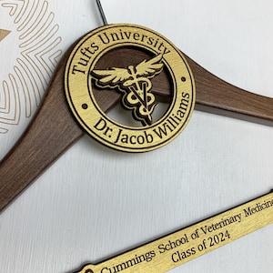 May include: A wooden hanger with a gold-colored round plaque that says "Tufts University Dr. Jacob Williams" with a caduceus symbol in the center. The hanger also has a gold-colored plaque that says "Cummings School of Veterinary Medicine Class of 2024".