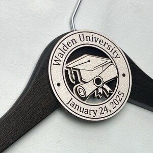 May include: A black wooden hanger with a white circular tag that says "Walden University" and "January 24, 2025". The tag also features a graduation cap and diploma.