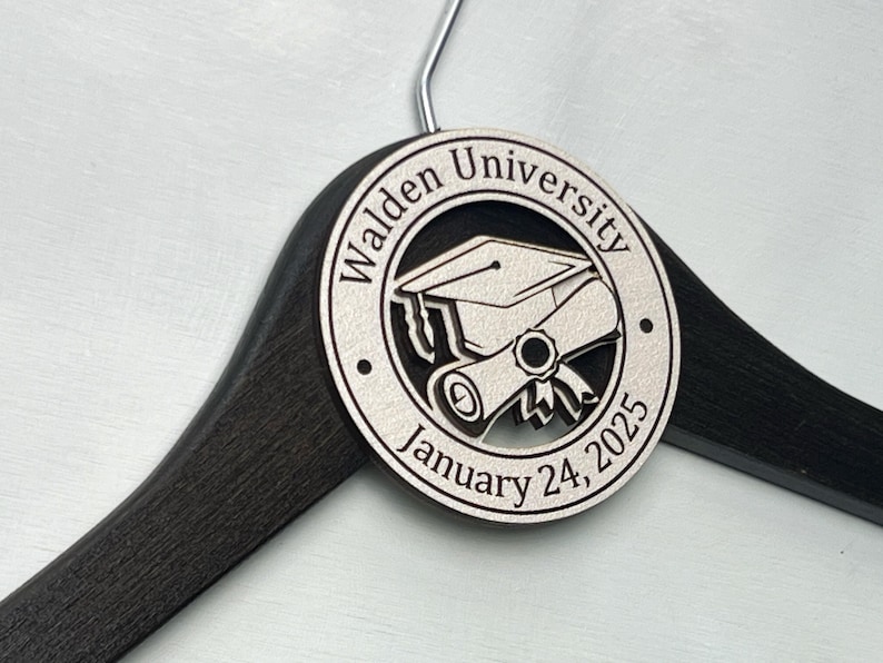 May include: A black wooden hanger with a white circular tag that says "Walden University" and "January 24, 2025". The tag also features a graduation cap and diploma.