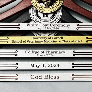 May include: A set of five engraved wooden hangers with silver and gold accents. The hangers are decorated with text that reads "White Coat Ceremony", "University of Cornell School of Veterinary Medicine Class of 2024", "College of Pharmacy Class of 2024", "May 4, 2024", and "God Bless".