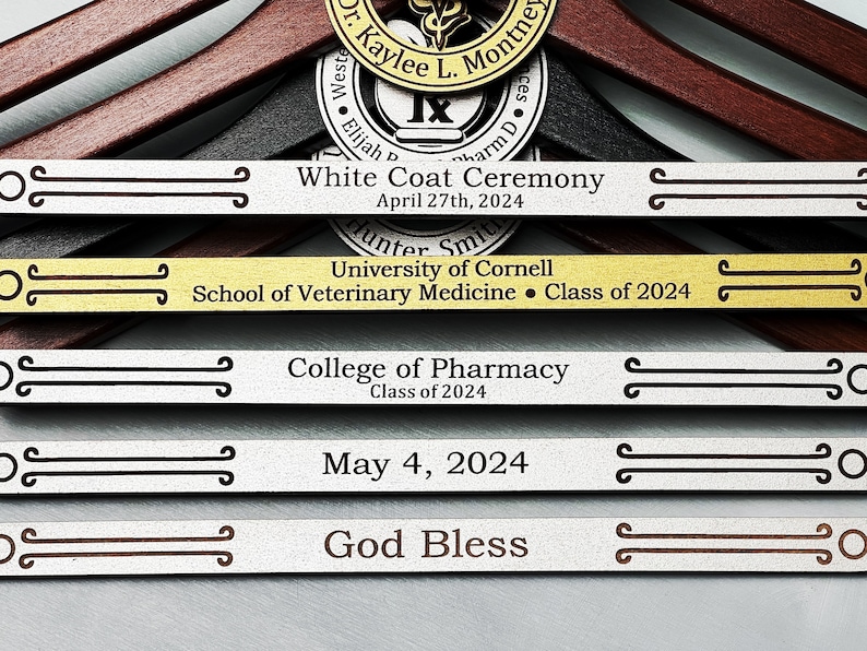 May include: A set of five engraved wooden hangers with silver and gold accents. The hangers are decorated with text that reads "White Coat Ceremony", "University of Cornell School of Veterinary Medicine Class of 2024", "College of Pharmacy Class of 2024", "May 4, 2024", and "God Bless".