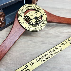 May include: A wooden hanger with a gold-colored round plaque that says "Walden University January 24, 2025" and a graduation cap and scroll design. A gold-colored plaque with the text "Dr. Brittany Hamilton Ph.D Clinical Psychology" is attached to the hanger.