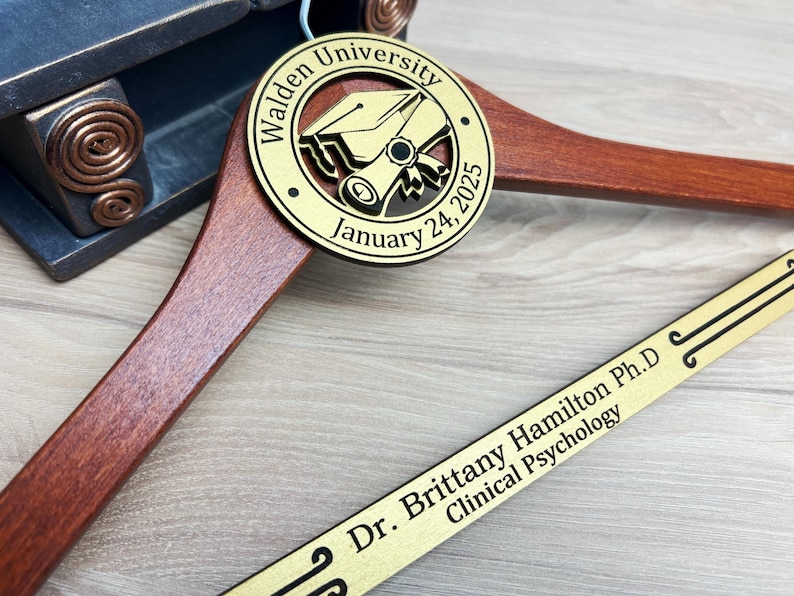 May include: A wooden hanger with a gold-colored round plaque that says "Walden University January 24, 2025" and a graduation cap and scroll design. A gold-colored plaque with the text "Dr. Brittany Hamilton Ph.D Clinical Psychology" is attached to the hanger.