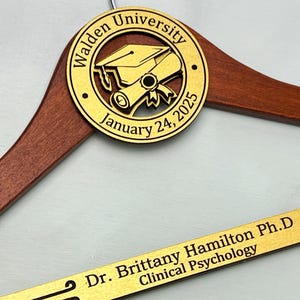 May include: A wooden hanger with a gold engraved circle featuring a graduation cap and scroll, the text "Walden University January 24, 2025", and a gold engraved nameplate with the text "Dr. Brittany Hamilton Ph.D Clinical Psychology".