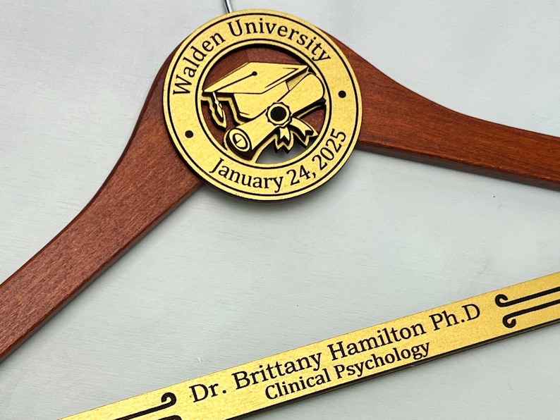 May include: A wooden hanger with a gold engraved circle featuring a graduation cap and scroll, the text "Walden University January 24, 2025", and a gold engraved nameplate with the text "Dr. Brittany Hamilton Ph.D Clinical Psychology".