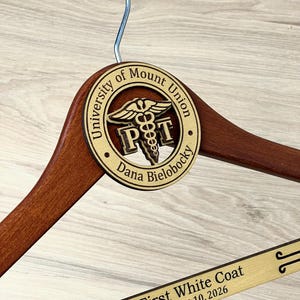 May include: A wooden clothes hanger with a silver metal hook. A circular wooden plaque features the text University of Mount Union, Dana Bielobocky, and a caduceus symbol with PT. The bottom bar is engraved with First White Coat August 10, 2026.