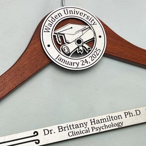 May include: A wooden hanger with a silver metal plaque featuring a graduation cap and scroll design. The plaque reads "Walden University January 24, 2025". A silver metal nameplate reads "Dr. Brittany Hamilton Ph.D. Clinical Psychology".