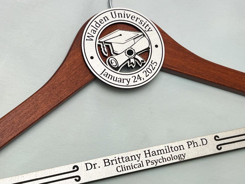 May include: A wooden hanger with a silver metal plaque featuring a graduation cap and scroll design. The plaque reads "Walden University January 24, 2025". A silver metal nameplate reads "Dr. Brittany Hamilton Ph.D. Clinical Psychology".