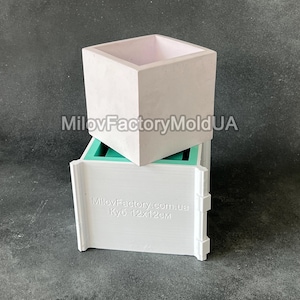 May include: A square, light gray concrete planter sits atop a white plastic mold. The planter has a light pink interior. The mold is white with the text "MilovFactory.com.ua" and "Куб 12x12cm".