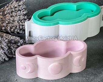 May include: A pale pink cloud-shaped container with moon and circle designs. A white and green cloud-shaped mould is behind it. The container is likely for decorative or functional use.