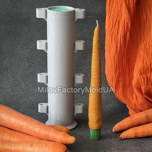 May include: An orange carrot-shaped candle with a green base is displayed beside a grey candle mould. Fresh carrots and orange fabric are also in the image. The mould is cylindrical with small, square protrusions.
