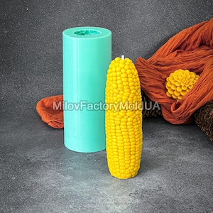 May include: A teal silicone mold and a yellow corn-shaped candle. The candle is sitting on a gray surface.