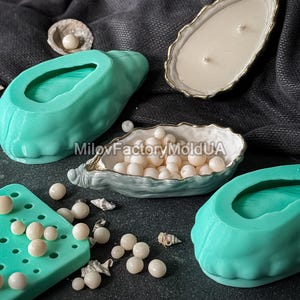 May include: An assortment of decorative items, including teal oyster-shaped molds, a candle in a gold-trimmed shell, and a tray filled with white pearls. Small seashells and scattered pearls add to the coastal theme, set against a dark background.