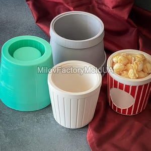 Peut inclure: Quatre récipients de formes et de couleurs différentes. Il y a un cylindre turquoise, un cylindre gris, un récipient blanc avec des lignes verticales et un récipient rayé rouge et blanc rempli de pop-corn. Le texte "MilovFactoryMoldUA" est visible.