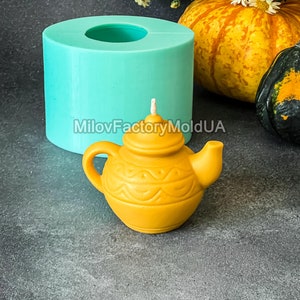 May include: A teal silicone mould for making a teapot-shaped candle. The mould is sitting on a grey surface next to a yellow teapot-shaped candle and a small pumpkin.