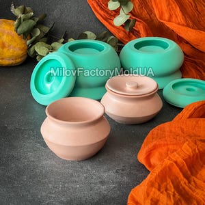 Può includere: Una collezione di stampi in ceramica di varie dimensioni e forme, tra cui vasi e ciotole rotonde, in tonalità di rosa e verde acqua. L'immagine presenta anche tessuto arancione e vegetazione.