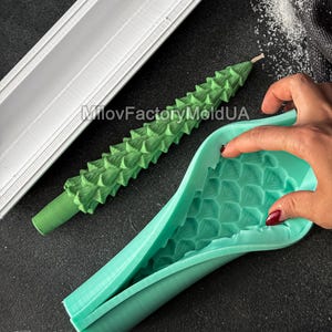 Puede incluir: Una vela verde con forma de árbol de Navidad junto a un molde de silicona turquesa y un molde de plástico blanco. La vela tiene una superficie texturizada con un diseño detallado de árbol. Los moldes están sobre una superficie oscura.