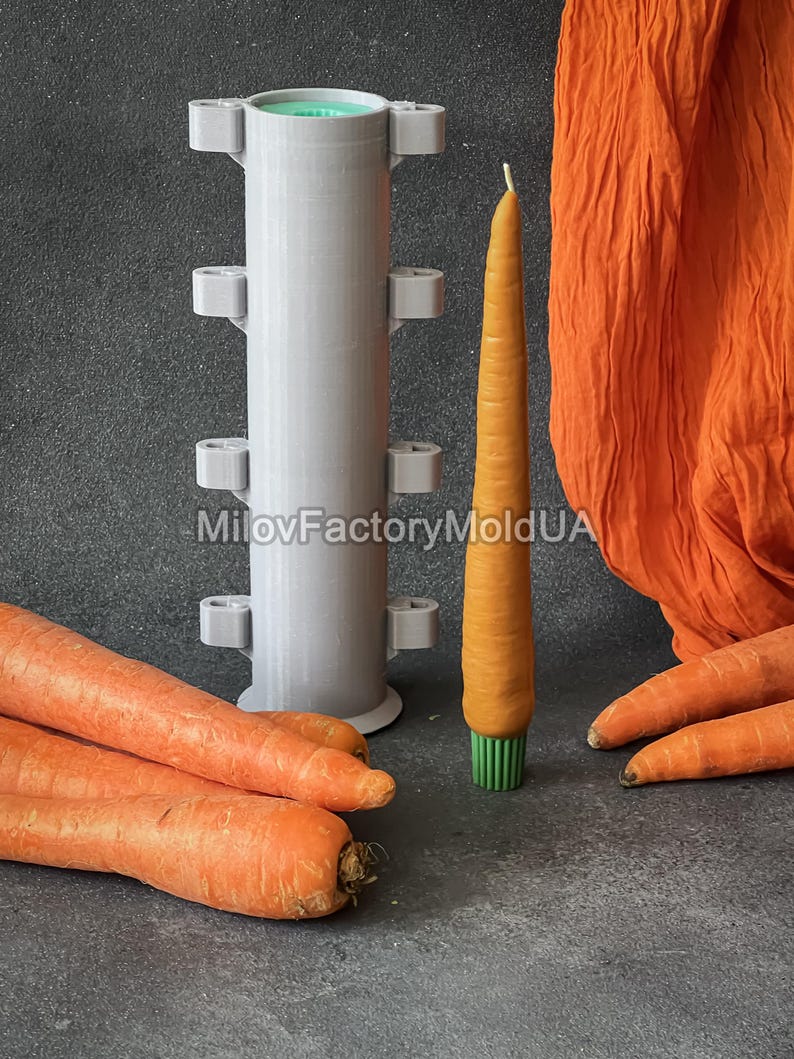 May include: An orange carrot-shaped candle with a green base is displayed beside a grey candle mould. Fresh carrots and orange fabric are also in the image. The mould is cylindrical with small, square protrusions.