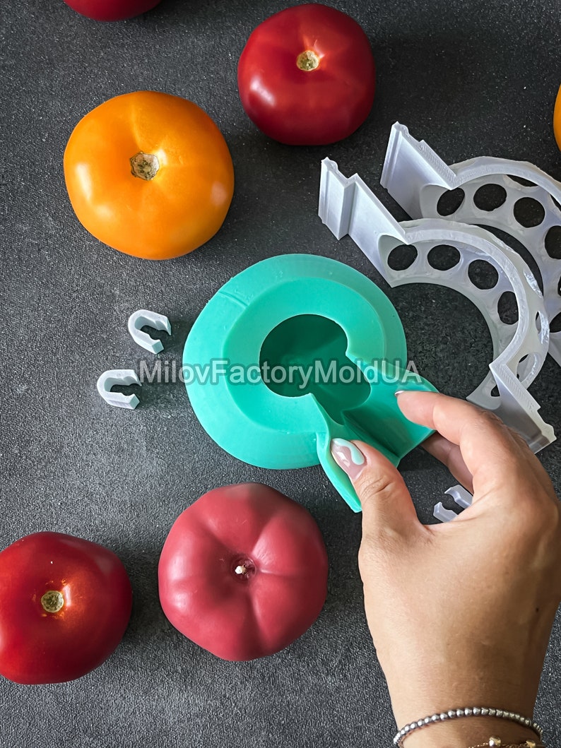 May include: A teal silicone mold shaped like a tomato, with a hand holding the mold open to reveal the inside. The mold is on a gray surface with several red and orange tomatoes and white plastic pieces.