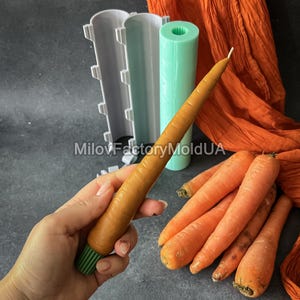 May include: A carrot-shaped candle held in a hand, alongside fresh carrots. The candle is orange with a green base. Candle moulds and orange fabric are in the background. The text "MilovFactoryMoldUA" is visible.