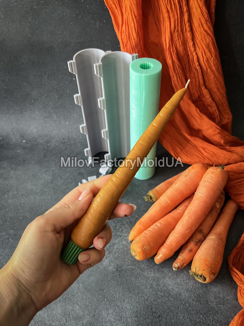 May include: A carrot-shaped candle held in a hand, alongside fresh carrots. The candle is orange with a green base. Candle moulds and orange fabric are in the background. The text "MilovFactoryMoldUA" is visible.