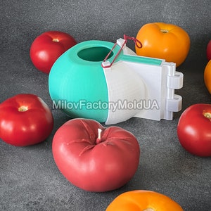 May include: A 3D printed mold for making heart-shaped tomatoes. The mold is white and teal with a red rubber band. There are several red and orange tomatoes around the mold. The text "MilovFactoryMoldUA" is printed on the mold.