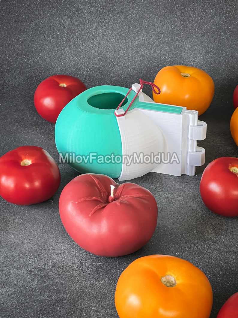 May include: A 3D printed mold for making heart-shaped tomatoes. The mold is white and teal with a red rubber band. There are several red and orange tomatoes around the mold. The text "MilovFactoryMoldUA" is printed on the mold.