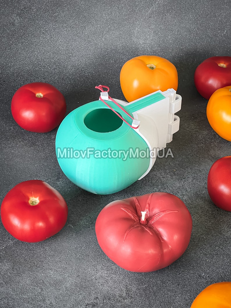 May include: A teal silicone mold shaped like a tomato, with a white plastic base and a red rubber band. The mold is surrounded by red and orange tomatoes.