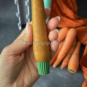 May include: An orange carrot-shaped silicone mould with a green base is held in a hand. The text "MilovFactoryMoldUA" is visible. Several real carrots are in the background. The mould is likely for making decorative items.