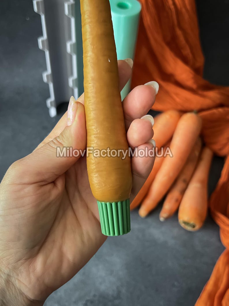 May include: An orange carrot-shaped silicone mould with a green base is held in a hand. The text "MilovFactoryMoldUA" is visible. Several real carrots are in the background. The mould is likely for making decorative items.