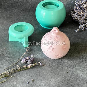 Pu&ograve; includere: Tre stampi in silicone per la realizzazione di candele. Gli stampi hanno la forma di un barattolo rotondo, un barattolo rotondo con coperchio e un ornamento rotondo con un motivo a fiocco di neve. Gli stampi sono su una superficie grigia.