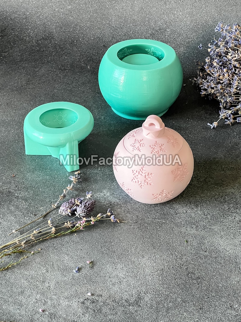 Pu&ograve; includere: Tre stampi in silicone per la realizzazione di candele. Gli stampi hanno la forma di un barattolo rotondo, un barattolo rotondo con coperchio e un ornamento rotondo con un motivo a fiocco di neve. Gli stampi sono su una superficie grigia.