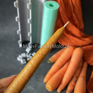 May include: A carrot-shaped candle with a white wick is held in front of a grey surface. Several orange carrots and candle moulds are also visible. The moulds are white and mint green. The background includes orange fabric.